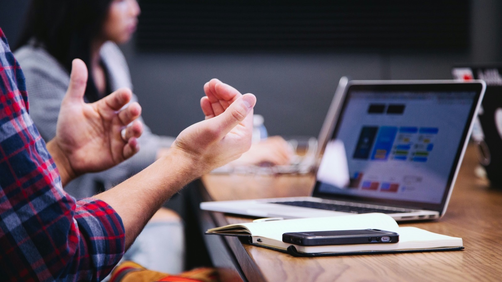 A person gesturing with both hands during a meeting, next to an open laptop and a notebook on a wooden table.