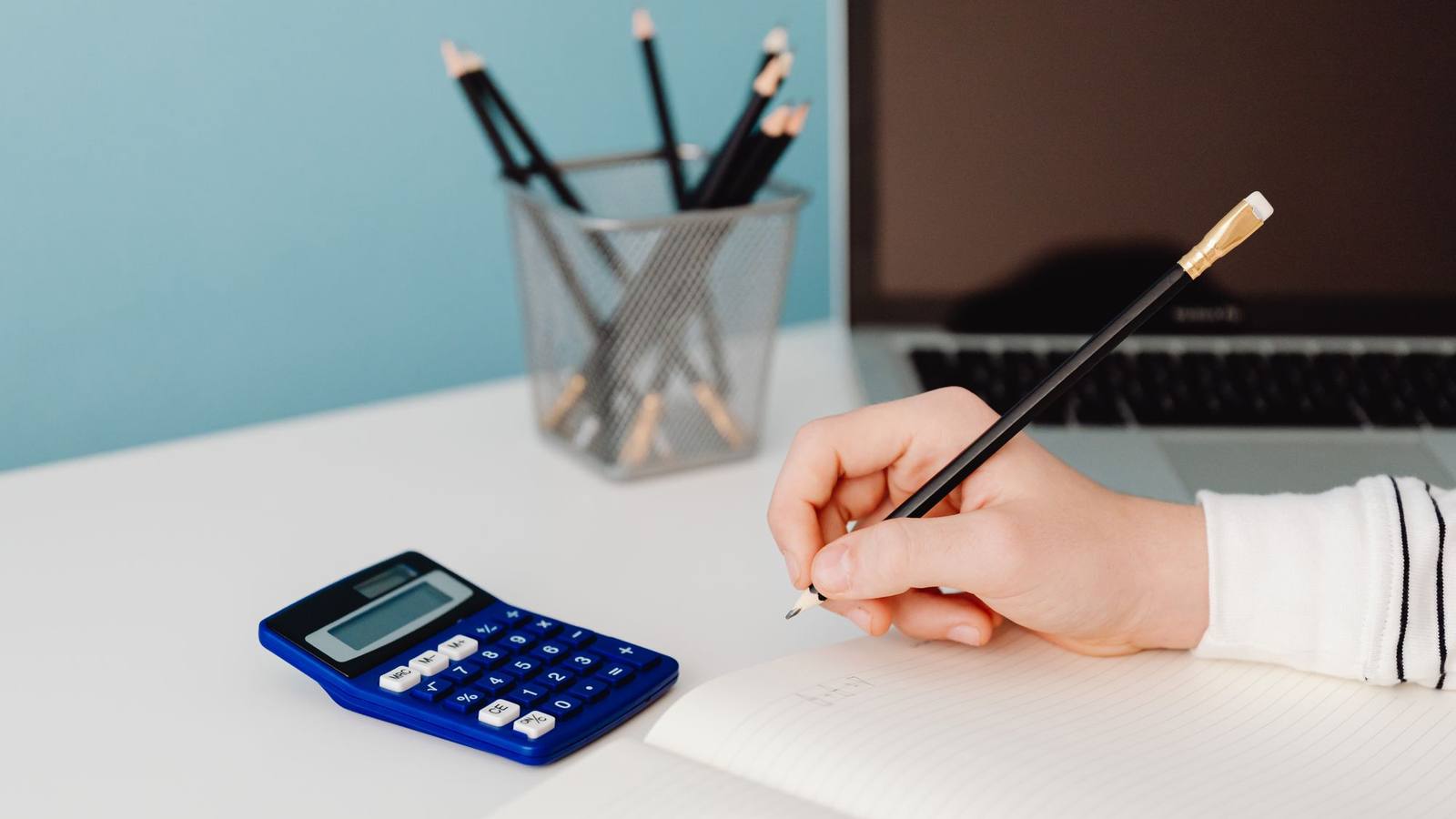 A hand writing in a notebook next to a calculator and a laptop.